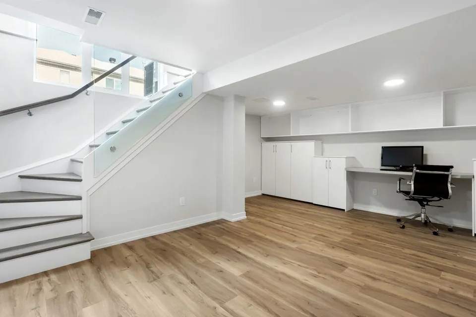 Bright finished basement room with a glass-balustrade staircase, high windows and a built-in desk